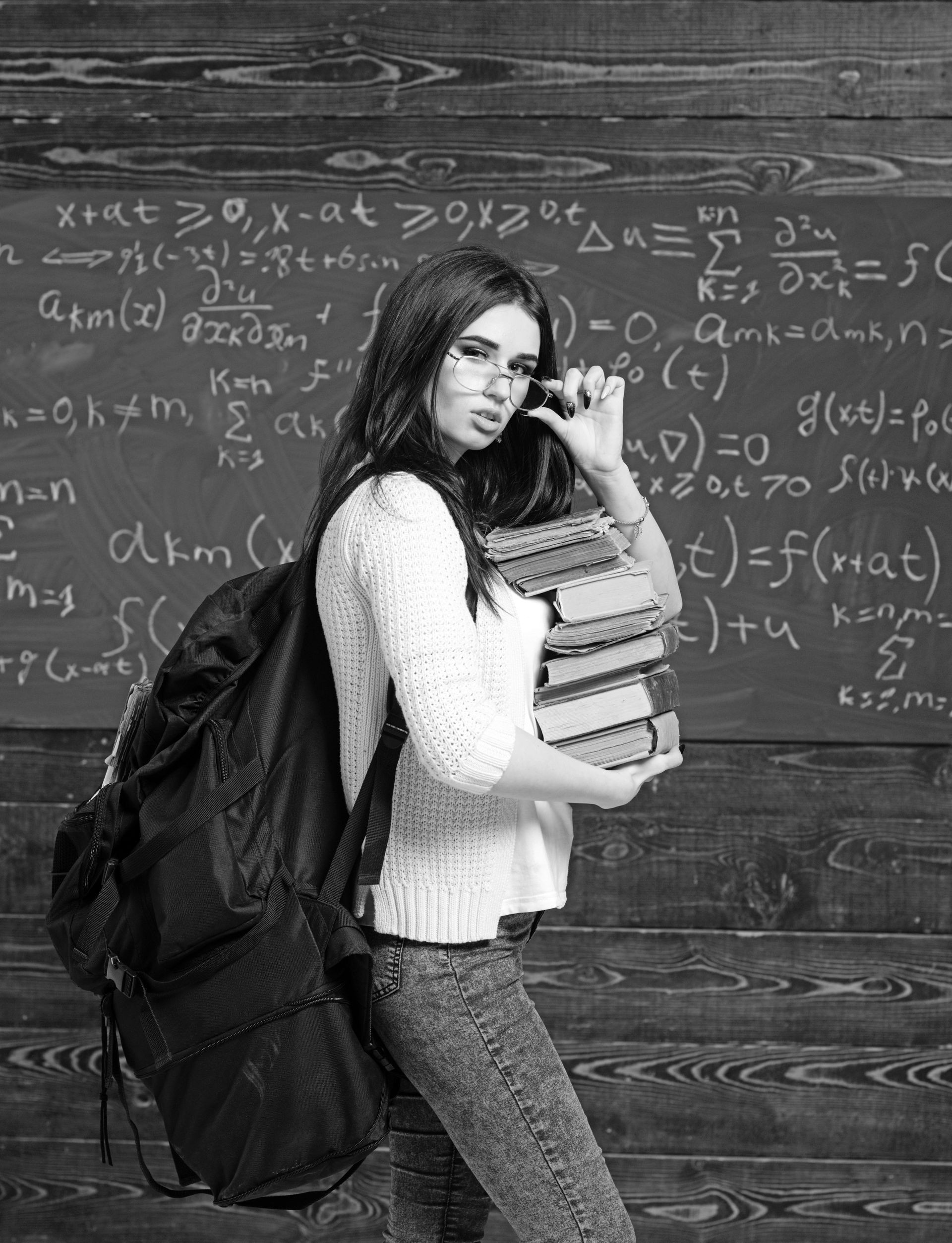 Young brunette student with heap of books and heavy rucksack holding her glasses. College education and pressure before final exams.